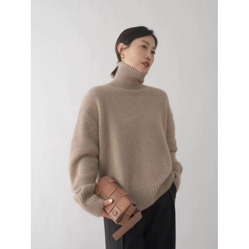 Woman wearing a beige turtleneck sweater holding a brown leather bag against a plain background