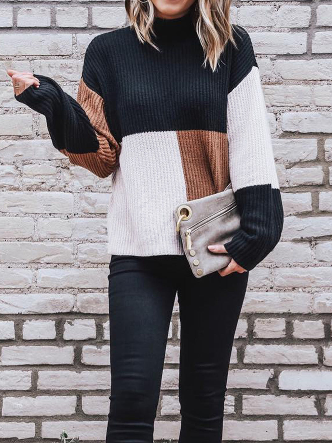 Person wearing a color-blocked sweater and holding a clutch against a brick wall.